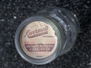Close-up photograph of a glass Rivermead Dairy bottle lying on its side. The paper cap reads “Rivermead Dairy, Cornwall” and identifies the contents as a chocolate-flavoured dairy drink. The bottle appears empty and is shown against a dark textured background.
