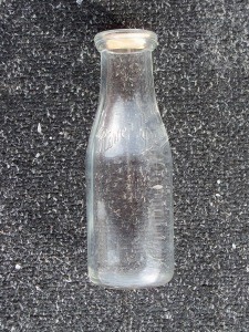 Photograph of a small, clear glass dairy bottle standing upright against a dark textured background. The bottle is empty, with visible scuffs and residue on the glass, and has a light-coloured cap or stopper at the top.