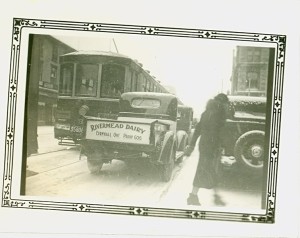 Black-and-white historic photograph of a Rivermead Dairy delivery truck on a city street. A streetcar is visible ahead of the truck, with tracks running along the roadway. A pedestrian in a dark coat walks beside parked vehicles on the right. The photograph is framed by a decorative printed border.