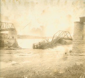 Sepia-toned historic photograph showing the aftermath of the Cornwall Bridge Disaster. A large metal truss bridge has partially collapsed into the river, with twisted bridge sections resting in the water between two surviving stone piers. Portions of the bridge remain standing on both sides of the river. Small groups of people are visible near the bridge approaches and along the shoreline, looking toward the wreckage.