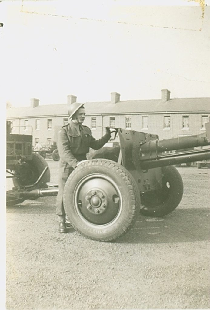 A soldier in uniform and helmet stands beside a large artillery gun in an open courtyard with brick buildings in the background. The mood is solemn.
