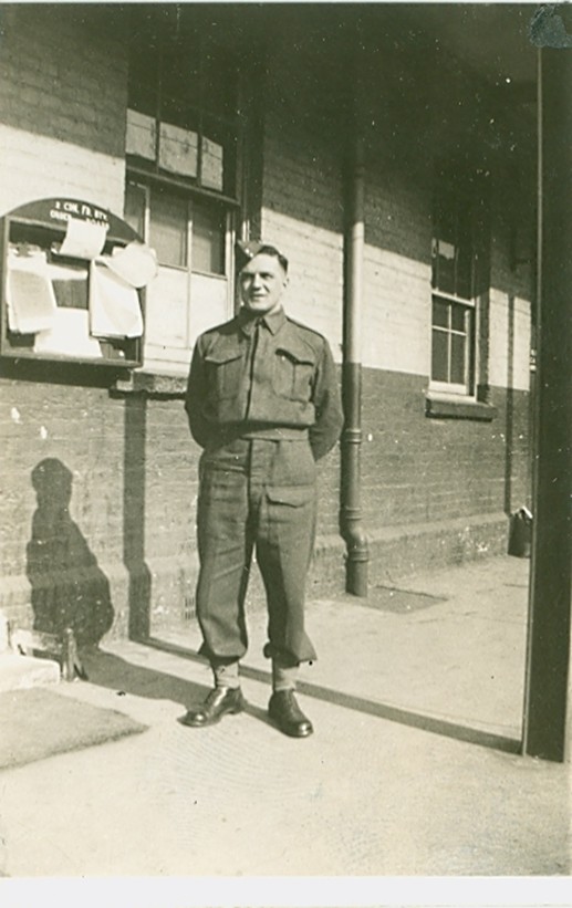 A man in a military uniform stands on a sunlit sidewalk beside a brick building. Papers are pinned to a nearby board, creating a nostalgic, historical feel.