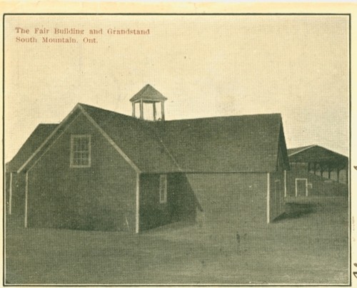 Alt text: Sepia-toned image of the Fair Building and Grandstand in South Mountain, Ontario. The structure is barn-like with a steeple, casting long shadows.
