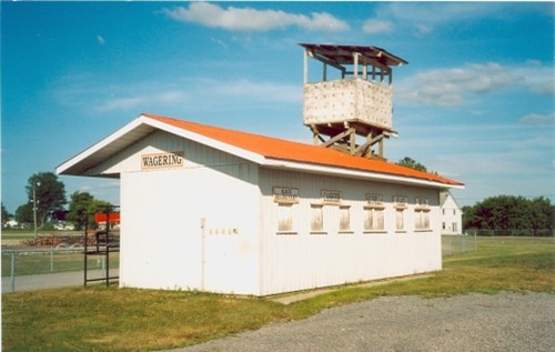 A small, white building with a red roof labeled "Wagering" under a sunny sky. A rustic wooden observation tower rises above it, surrounded by green grass.