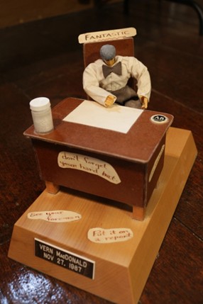 A small desk figurine with a person sitting in a chair labeled "Fantastic." The desk has a coffee cup and paper, with humorous notes about leaving early and mistakes.