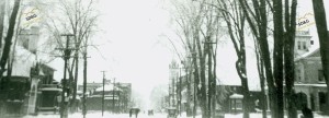 A snowy street scene with bare trees lining the road. Buildings and utility poles are visible. People in coats walk in the distance, conveying a cold, tranquil atmosphere.