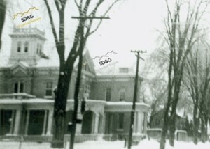 A snowy street scene depicts a historic, large brick house with a tower and porch, surrounded by bare trees and telephone poles in winter.