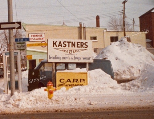Snowy street scene with "KASTNERS" menswear sign and large snow pile. A yellow fire hydrant is visible. Buildings and additional signs in the background.