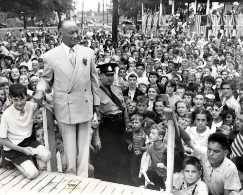 A man in a suit stands on a platform with a police officer, surrounded by a large, diverse crowd of children and adults, smiling and attentive. Festive atmosphere.