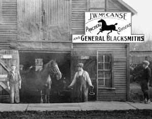 Vintage photo of a blacksmith shop with a sign reading "J.W. McCanse Practical Shoers and General Blacksmiths." A horse stands outside, held by a worker. Two men watch nearby.
