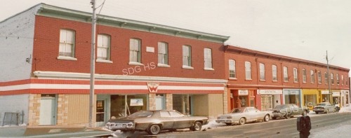 Street view of a retro row of brick shops with vintage cars parked along the sidewalk. The scene has a nostalgic, small-town feel on a cloudy day.