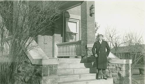 A man in a long coat and hat stands on the steps of a brick house. Leafless trees frame the scene, conveying a winter setting and calm mood.