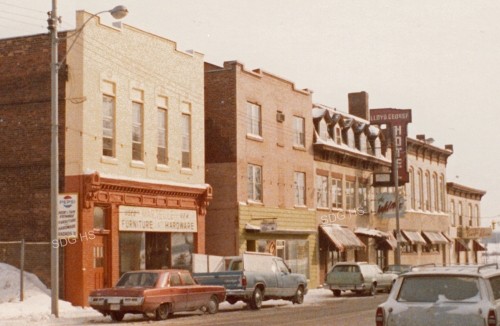 A row of vintage brick buildings line a snowy street, featuring a furniture and hardware store, parked cars, and a hotel sign. The scene feels nostalgic.