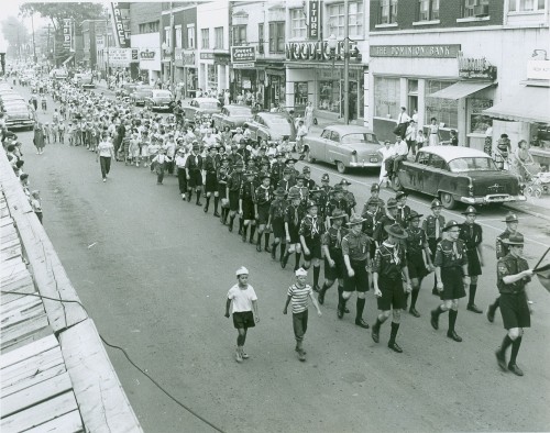 A vintage parade scene with marching bands and scouts walking down a street lined with 1950s cars and shops, creating a festive and nostalgic atmosphere.