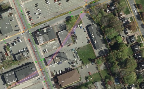 Aerial view of an urban intersection with marked roads and aerial lines, surrounded by buildings, a parking lot, and green spaces, conveying a detailed city layout.