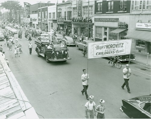 A lively street parade with people walking alongside a fire truck. Participants hold a banner for "Mayer Horovitz Annual Children's Picnic." Energetic and festive atmosphere.