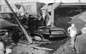 A coal carrying train car has overturned, spilling its load onto a crushed car. Bystanders, including children, gather around, observing the chaotic scene.