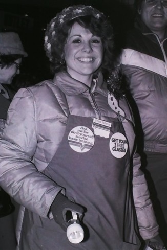 A smiling woman in a winter coat and gloves stands in a crowd, holding a small object. Her coat has several buttons with messages on it. The mood is lively.