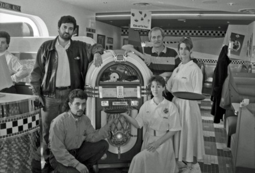 A group of five people, three men and two women, pose with a vintage jukebox in a retro diner. The setting has a nostalgic 1950s theme.
