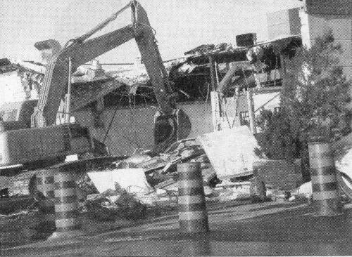 A large excavator demolishes an industrial building, surrounded by debris and caution barrels, conveying a sense of destruction and construction.