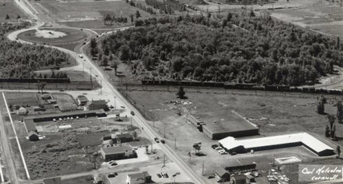 Aerial view of a rural town with a main road, scattered buildings, and a train passing through. Surrounding areas feature dense trees and open fields. The mood is serene.