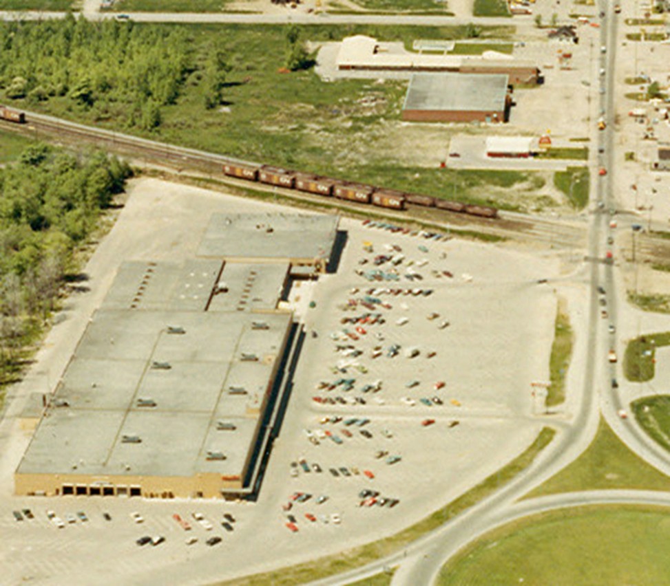 Aerial view of a large industrial building with a busy parking lot filled with cars. A train passes nearby on tracks lined with green trees, set in an urban area.