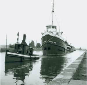 A large ship, labeled "South Wind," navigates a narrow canal, assisted by a small tugboat on its left. The scene conveys a sense of teamwork.