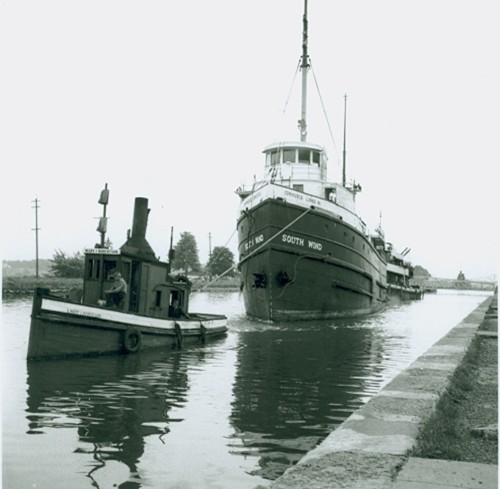 A large ship, labeled "South Wind," navigates a narrow canal, assisted by a small tugboat on its left. The scene conveys a sense of teamwork.