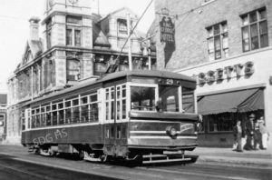 A vintage streetcar marked "Hastings East" travels down a street past early 20th-century buildings, including the historic St. George Hotel. Passersby watch it go by, evoking nostalgia.