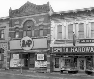 Historic black and white photo of A&P Grocery and Smith Hardware stores on a street. Brick buildings with vintage signage create a nostalgic atmosphere.
