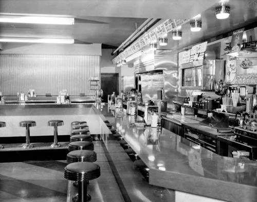 Black and white photo of a vintage diner with empty chrome stools along a counter. The decor is retro with a checkerboard floor, creating a nostalgic feel.