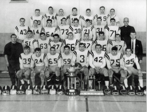 A vintage black and white photo of a football team and coaches. Players wear numbered jerseys and helmets are displayed. Trophies are in front, conveying achievement.