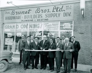 A group of men in suits stand in front of Brunet Bros. Ltd. hardware store, smiling and holding a ribbon for a grand opening. The building displays large signage.