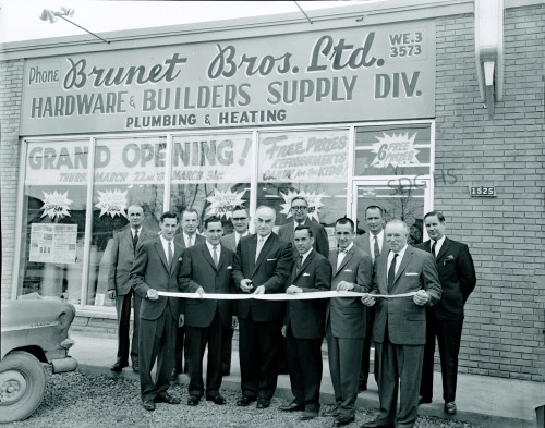 A group of men in suits stand in front of Brunet Bros. Ltd. hardware store, smiling and holding a ribbon for a grand opening. The building displays large signage.