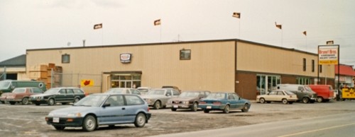 A large beige automotive service shop with several parked cars in front. Flags adorn the roof, and a sign is visible on the side, creating a retro vibe.