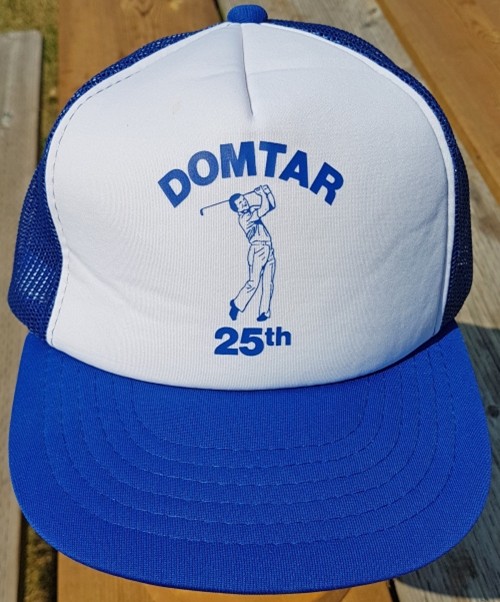 Blue and white trucker hat with "DOMTAR 25th" and a golfer illustration. The cap rests on a wooden surface, conveying a casual sports vibe.