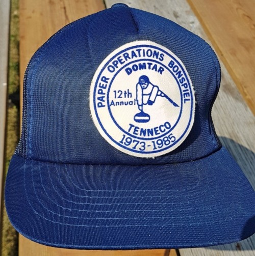 Blue cap featuring a circular patch with a curling graphic. Text reads "Paper Operations Bonspiel, 12th Annual, Domtar, Tenneco, 1973-1985."