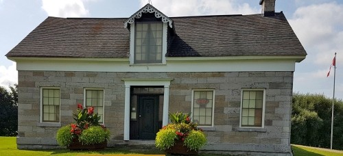 A quaint, stone house with a steep gabled roof and white trim sits in a grassy area. Flower boxes and a Canadian flag add charm under a blue sky.