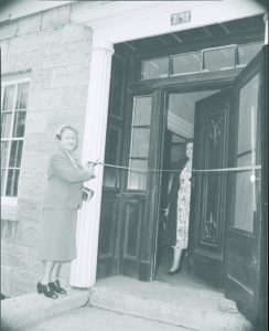 A woman in a suit cuts a ribbon at the entrance of a stone building, smiling slightly. Another woman stands in a floral dress inside, holding the door.