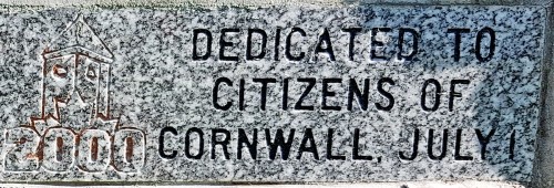 Stone plaque with an etched tower and "2000" icon, text reads "Dedicated to Citizens of Cornwall, July 1." Grey granite texture, sunlight casting shadows.