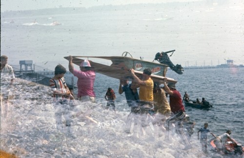 A group of people lift a wooden speedboat labeled "Miss Thriftway" onto a rocky shore by a large body of water. The scene is lively and collaborative.