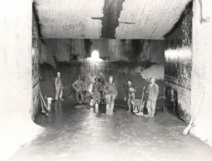 A group of workers stands in a large, dimly lit industrial chamber with concrete walls. Some hold tools, and a small window lets in light, creating a gritty, determined atmosphere.