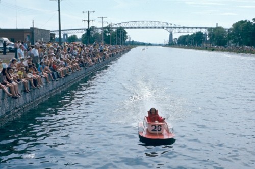 A person rides a small red speedboat numbered 26 through a canal, splashing water. A large crowd watches from the side. A bridge is visible in the background.