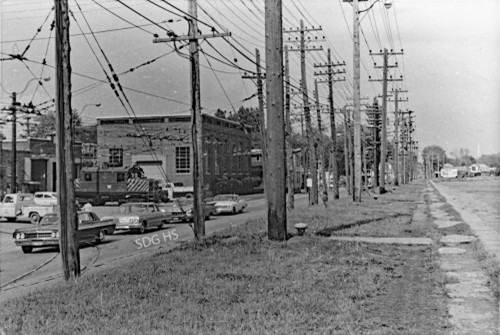 Black and white vintage street scene with utility poles lining the road. Classic cars drive past industrial buildings under a cloudy sky, conveying a nostalgic tone.