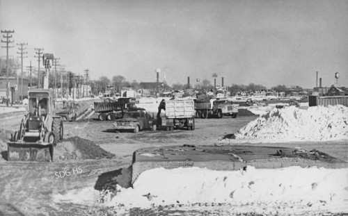 A snowy industrial scene with trucks and a bulldozer on a construction site. Chimneys and utility poles are visible in the background under a cloudy sky.