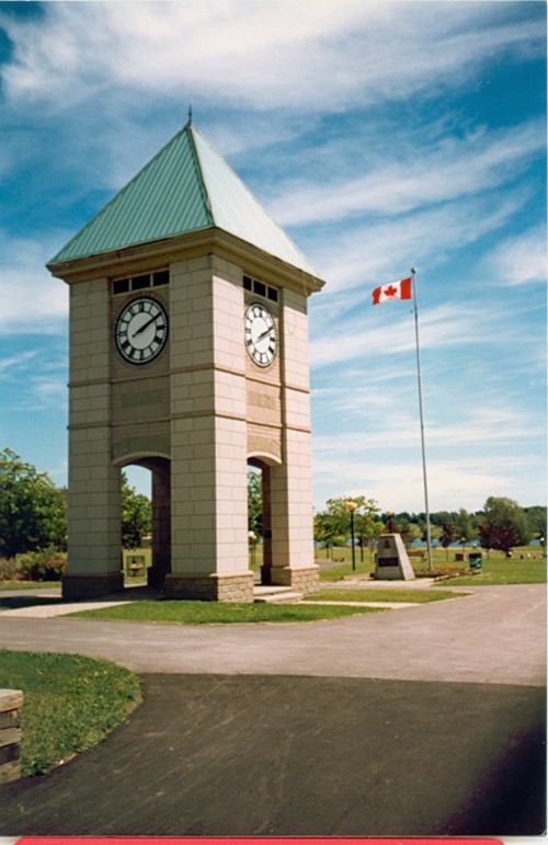 Clock tower with a green roof stands in a park under a bright blue sky. A Canadian flag waves on a pole nearby, creating a peaceful, patriotic scene.