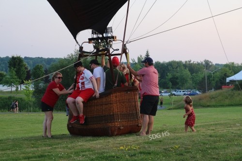 A group of people assist passengers onto a deflated hot air balloon basket on a grassy field. A child in red nearby, with trees and a tent in the background.