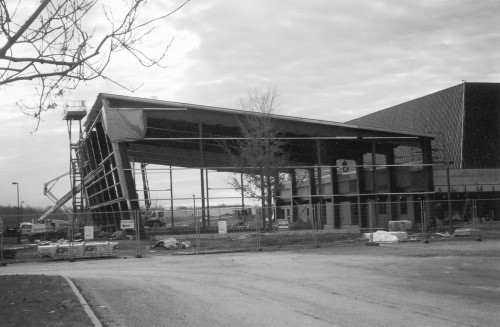 Steel framework of a large building under construction, surrounded by a chain-link fence. Construction materials and equipment are scattered around. Cloudy sky.
