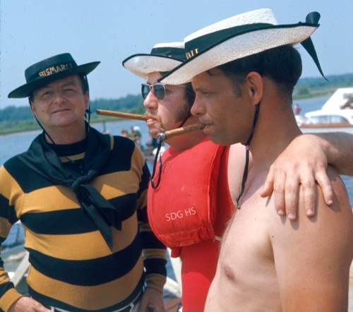 Three men on a boat deck wear sailor hats, with one in a striped shirt, another with a life vest and cigar, and a shirtless man. They appear relaxed and cheerful.