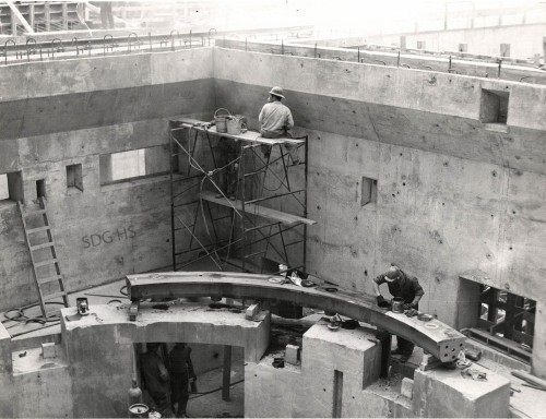 Construction site with workers installing large metal beams inside a deep concrete structure. Scaffolding and ladders visible, with a focused, industrious tone.